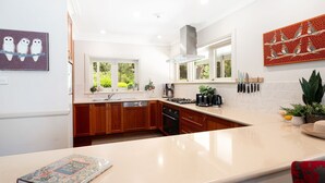 A modern kitchen with wooden cabinets, a white countertop, and a range hood. There are windows with views of greenery outside, and a decorative wall piece above the counter.