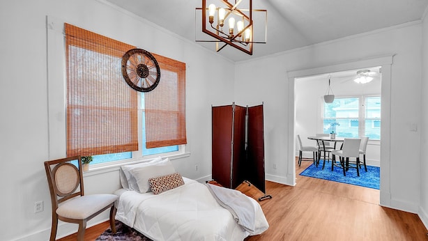 A bedroom with a bed, a chair, a window with blinds, a ceiling light fixture, and a view into a dining area. - C.a.l.m. Cottage At The Last Mile 2 Bedroom Home (Savannah)