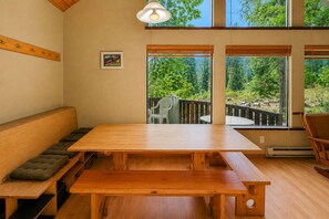 A wooden dining table with benches, a bench with a cushion, a hanging light fixture, and a view of a garden through large windows. - Icicle Chalet 1 Bedroom Cabin (Leavenworth)