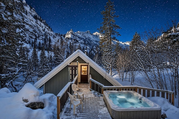 Un chalet en bois doté d’un bain à remous, entouré d’arbres et de montagnes enneigés, sous un ciel étoilé.