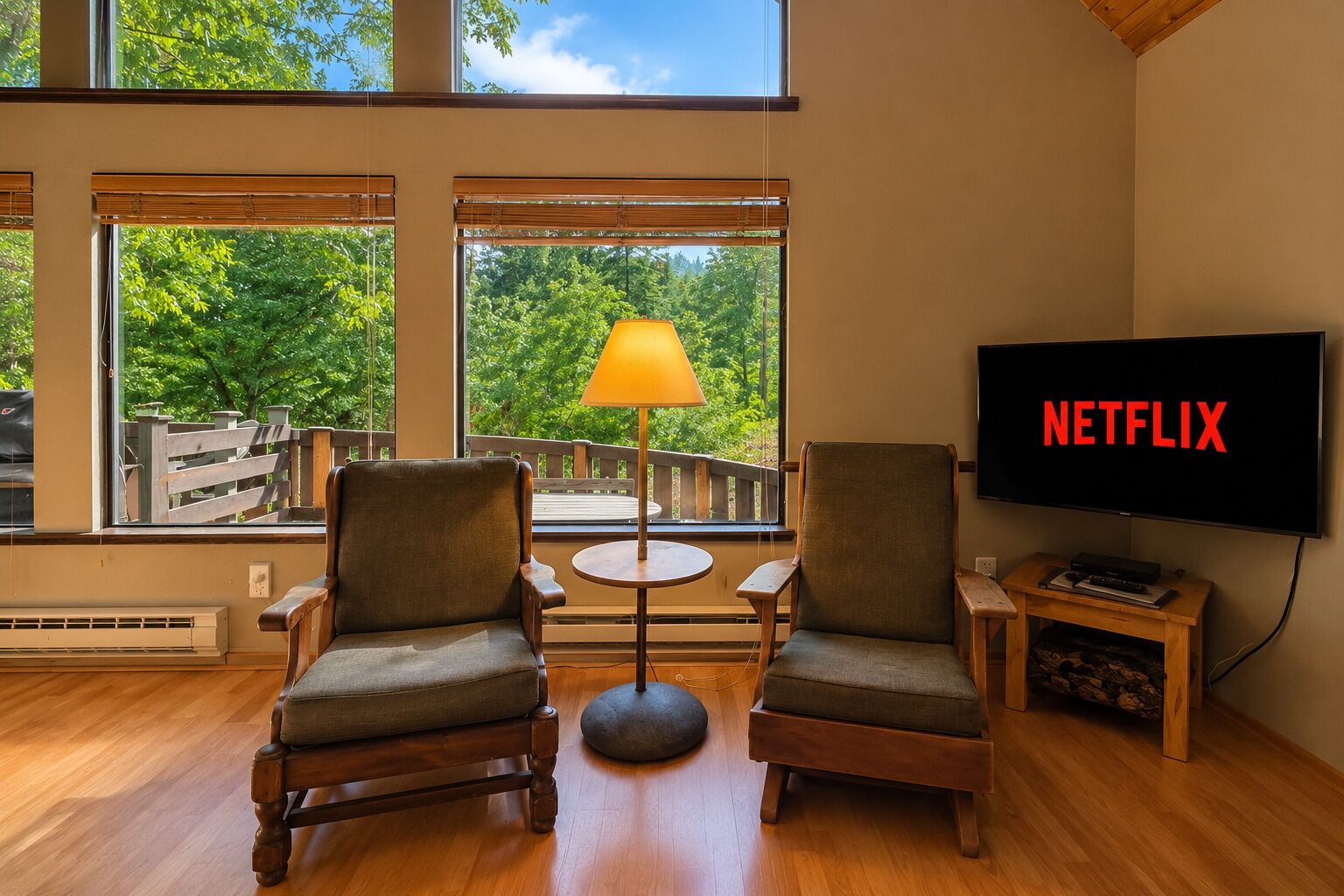 Quarto com duas cadeiras, uma mesa, uma luminária, uma TV exibindo 'NETFLIX' e amplas janelas com vista para árvores.