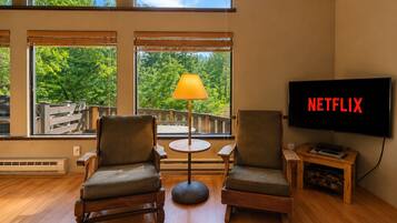 Quarto com duas cadeiras, uma mesa, uma luminária, uma TV exibindo 'NETFLIX' e amplas janelas com vista para árvores.