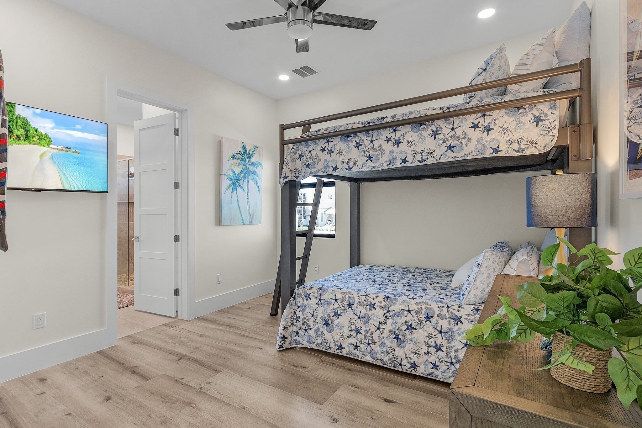 Ein Etagenbett mit Blumenmusterdecke, ein Deckenventilator, ein an der Wand befestigter Fernseher und ein Fenster mit Blick auf Palmen.