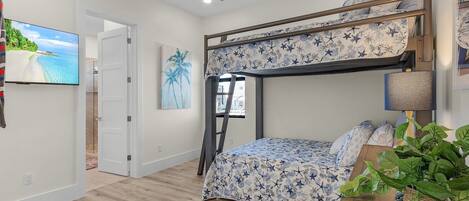 Ein Etagenbett mit Blumenmusterdecke, ein Deckenventilator, ein an der Wand befestigter Fernseher und ein Fenster mit Blick auf Palmen.