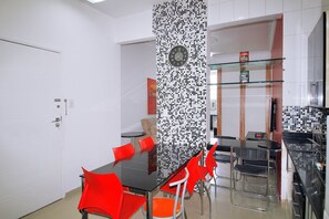 Un coin repas avec une table noire et des chaises rouges, une horloge au mur, et un espace cuisine comprenant un réfrigérateur et des étagères.