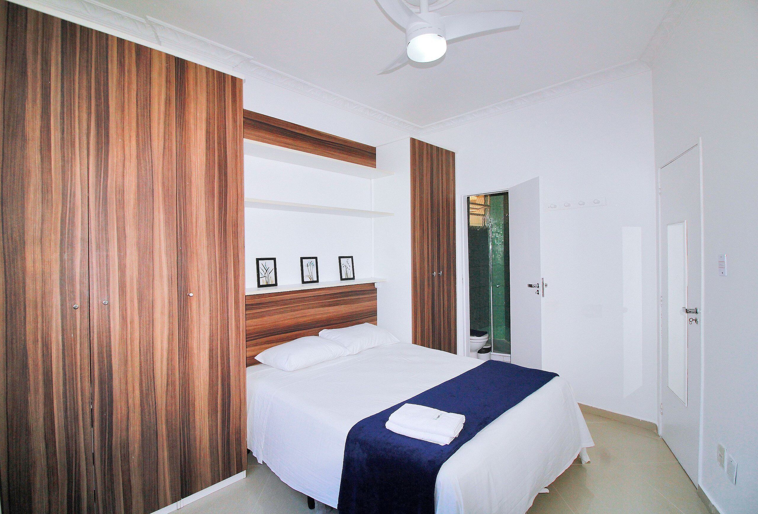 A neatly arranged bedroom with a bed, wooden wardrobe, and a ceiling fan.