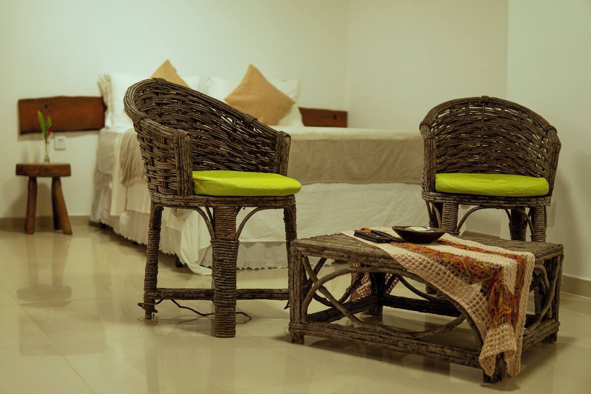 A room with two wicker chairs with green cushions, a small table with a bowl, and a bed with white bedding.