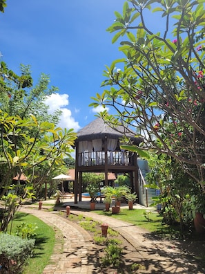 Relax in a serene garden setting with a shaded gazebo and brick pathway, surrounded by lush plants and a glimpse of a nearby pool.