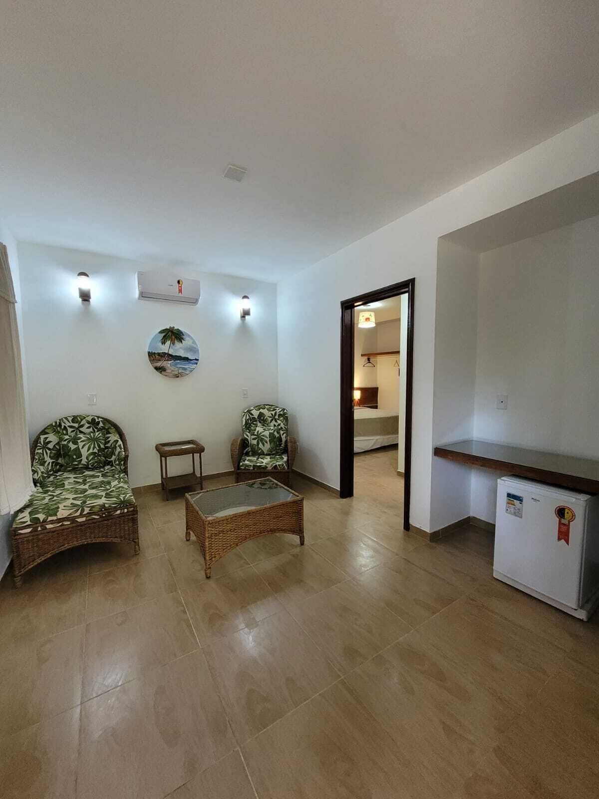 Una sala de estar con dos sillones de mimbre, una mesa de centro de vidrio, una mesita auxiliar y un refrigerador blanco.