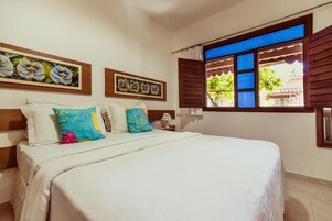 Un dormitorio con cama, almohadas, una mesita de noche y una ventana con contraventanas.