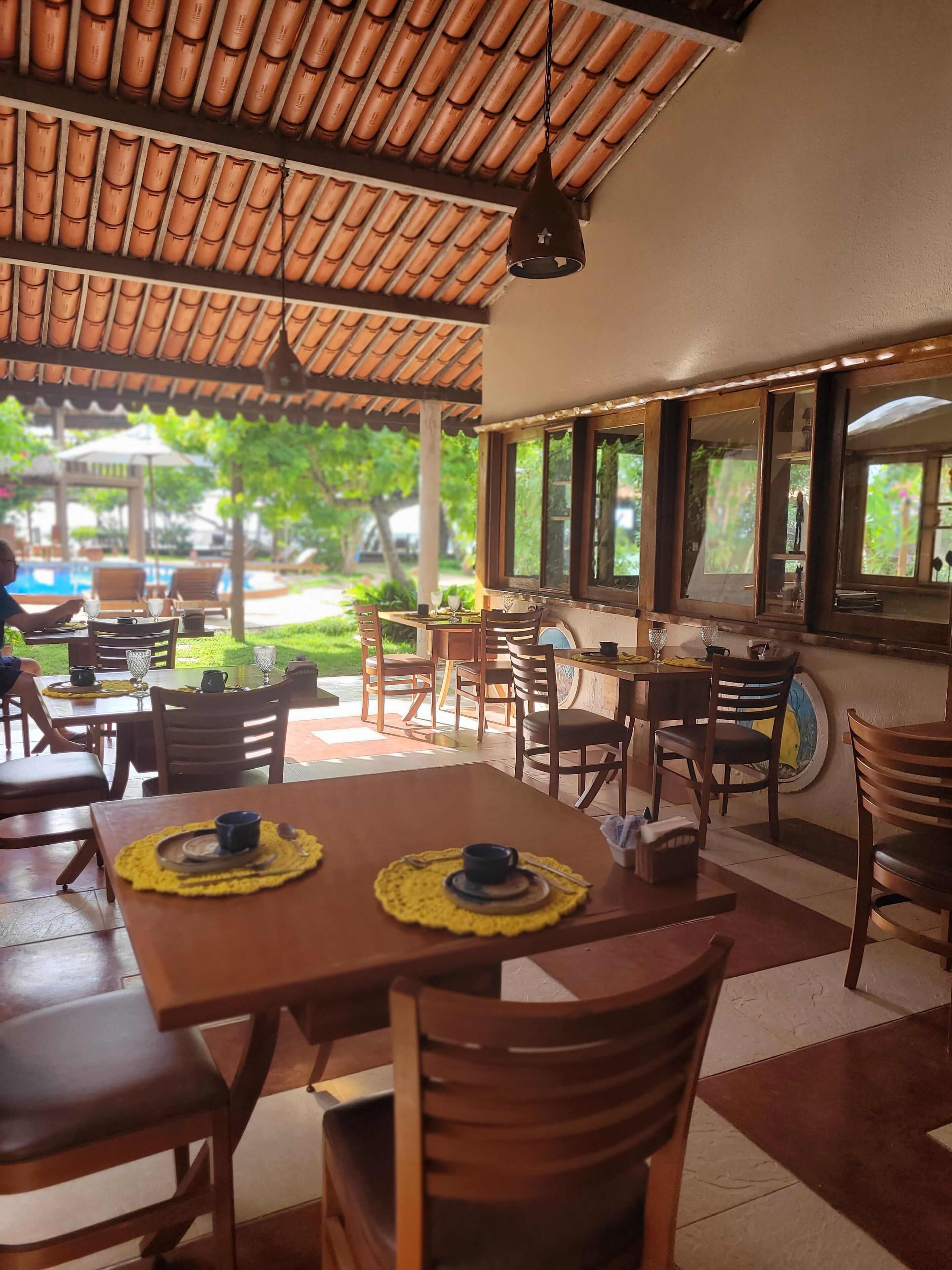 An outdoor cafe with wooden tables and chairs, a tiled roof, and a view of a swimming pool and greenery.