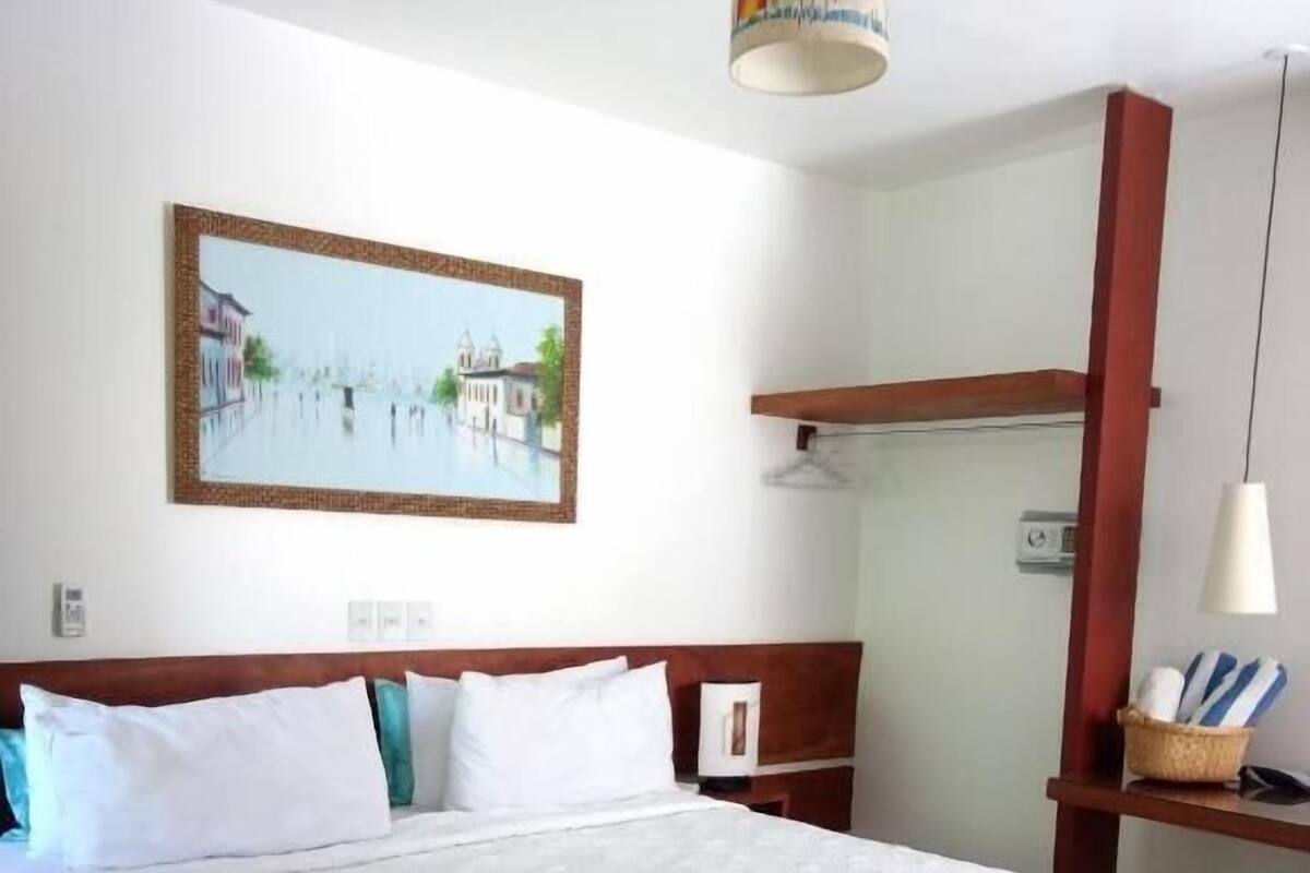 A hotel room with a large bed, a painting of a waterfront scene, a wooden headboard, and a small desk with a lamp.
