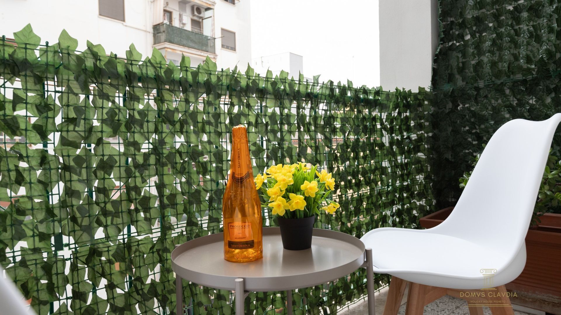 Ein Balkon mit weißem Stuhl, rundem Tisch, einer Flasche und einem Blumentopf mit gelben Blüten.