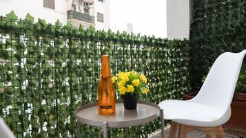 Ein Balkon mit weißem Stuhl, rundem Tisch, einer Flasche und einem Blumentopf mit gelben Blüten.