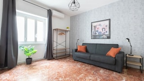 Un salon moderne avec un canapé gris, une plante, une étagère en métal et un tableau abstrait encadré accroché au mur.