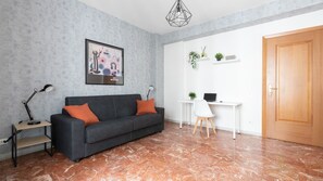 Un salon moderne avec un canapé foncé, une table d’appoint avec une lampe, une œuvre d’art abstraite encadrée, un bureau blanc avec un ordinateur portable et une porte en bois.