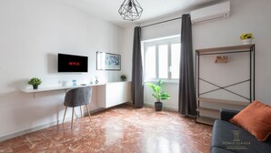 A modern living room with a TV, a desk, a chair, a couch, a plant, and a window with curtains. - Domvs Clavdia - Metro Station, 8 min to center, A-C (Roma)
