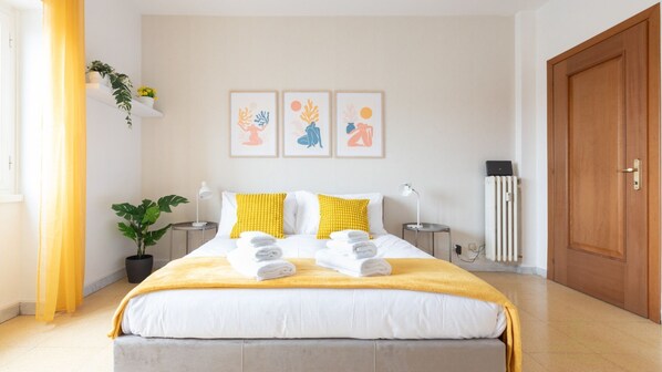 Une chambre d’hôtel avec un lit, deux tables de chevet, une plante en pot, un radiateur et une porte.