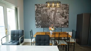 A dining area with a wooden table and chairs, a cityscape artwork on the wall, and a refrigerator in the background.