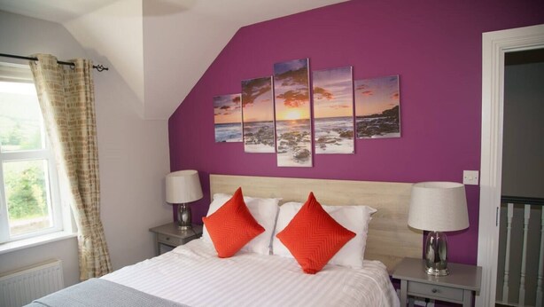 A bedroom with a bed, two nightstands, a window with curtains, and a purple wall with three landscape paintings.