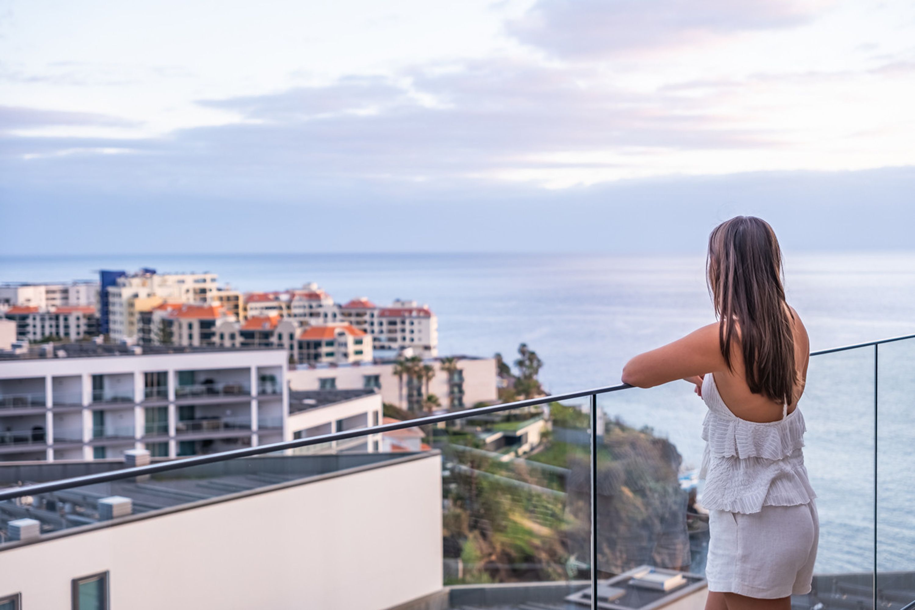 A woman standing on a balcony overlooking a coastal cityscape with buildings and the sea.