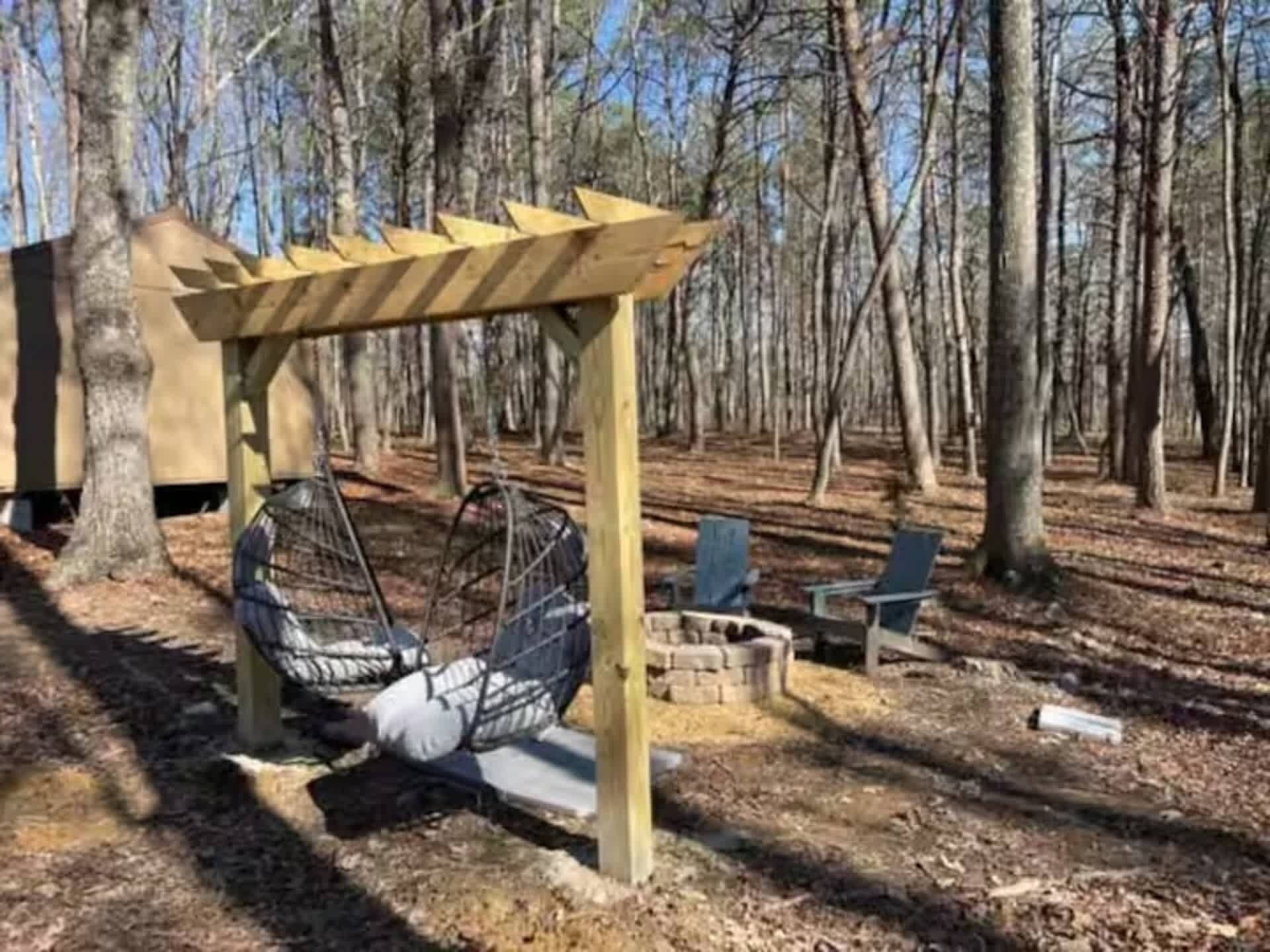 A wooden pergola with a swing chair and a fire pit in a wooded area.