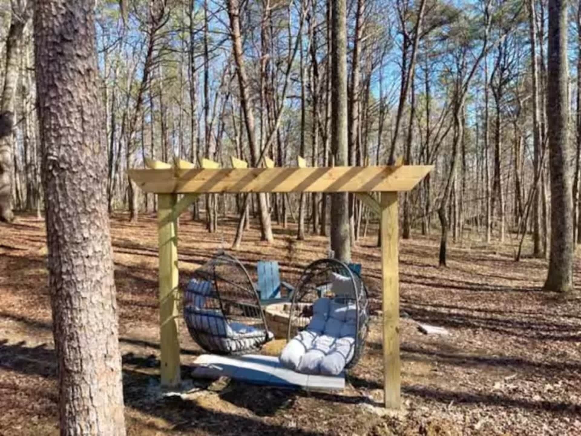 A wooden pergola with a swing chair and a cushioned bench in a forest setting.