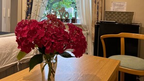 Una mesa de madera con un jarrón de flores rojas, una silla, un escritorio con una computadora y una ventana con cortinas.