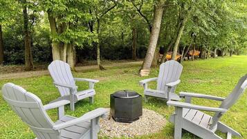 A circular arrangement of Adirondack chairs around a central fire pit in a wooded park setting.