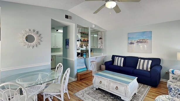 Uma sala de estar com sofá azul, cadeiras brancas, mesa de vidro, ventoinha no teto e um quadro com uma cena de praia.