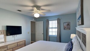 Um quarto com cama, televisão, ventoinha no teto, janela com cortinas e porta.