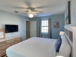 Ein Schlafzimmer mit Bett, Fernseher, Deckenventilator, einem Fenster mit Vorhängen und einer Tür.