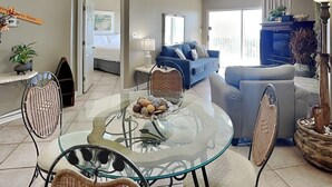 A modern living room with a glass dining table, wicker chairs, a sofa, and a ceiling fan. - Tidewater (Orange Beach) #108 | Gulf Front + Resort Amenities (Orange Beach)