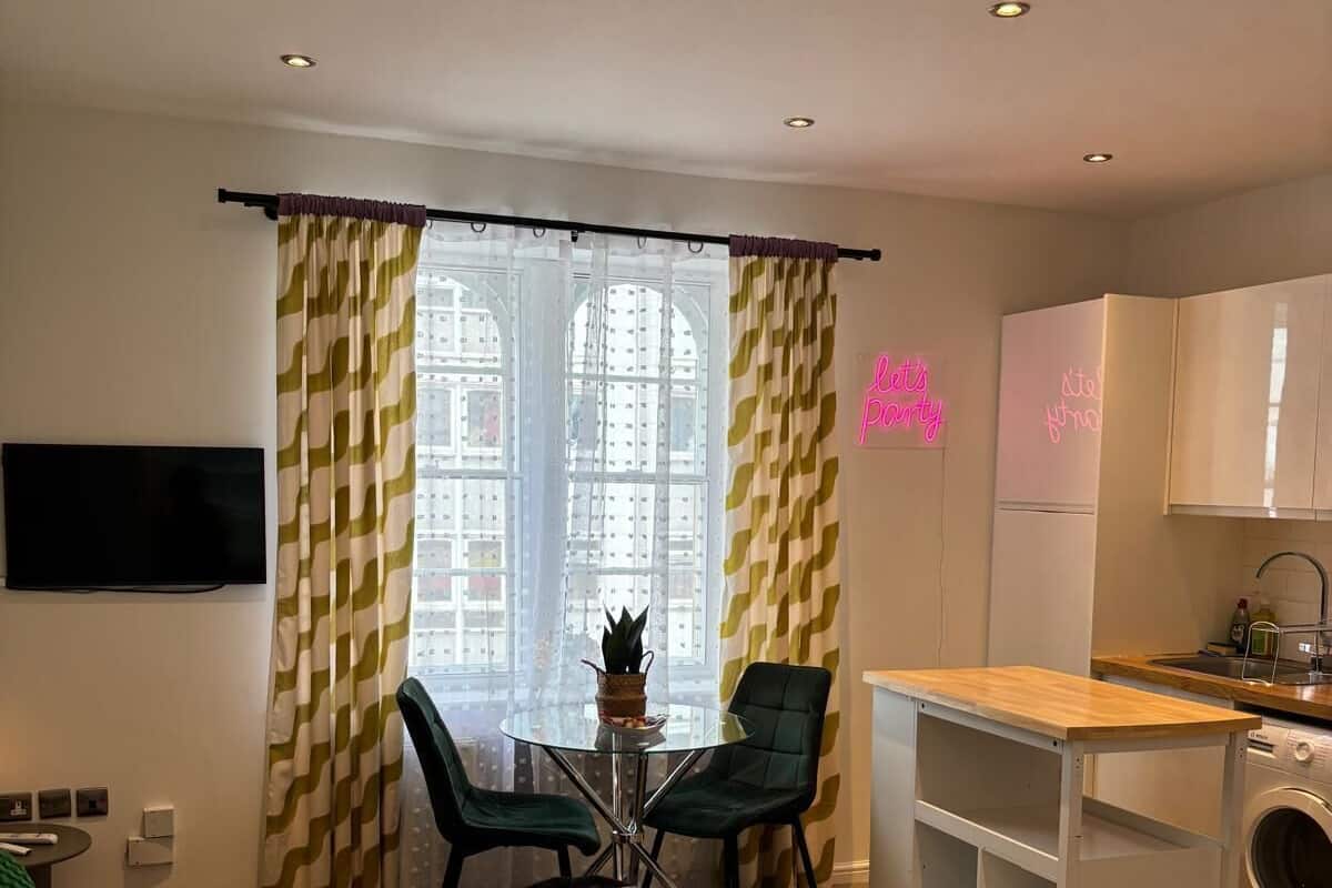 A compact hotel room with a bed, a small dining table with chairs, a washing machine, and a TV mounted on the wall.