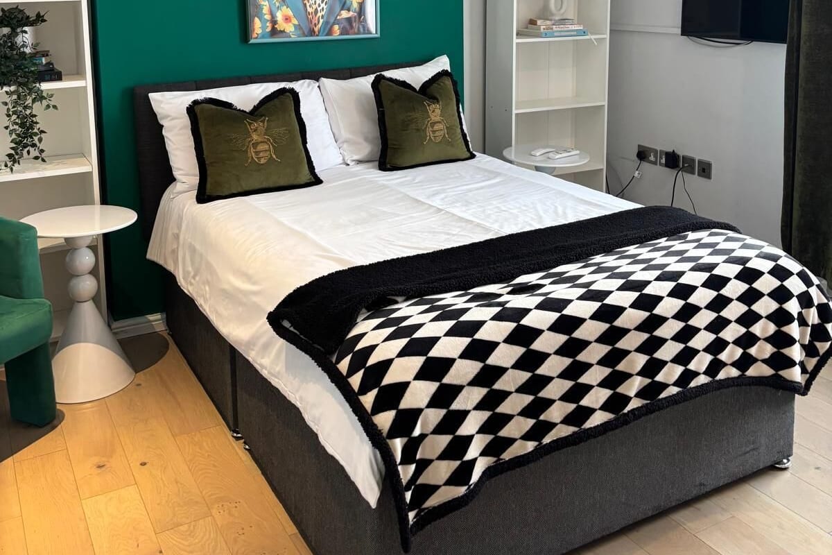A modern bedroom with a teal accent wall, a large bed with a checkered throw, a painting above the bed, and a TV mounted on the wall.