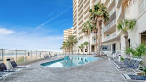A rooftop pool area with lounge chairs and palm trees, surrounded by a high-rise building. - Admiral's Quarters #1406 | Gulf Front + Amenities (Orange Beach)