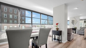A modern living room with a large window, a dining area with two chairs, a sofa, and a decorative mirror on the wall.