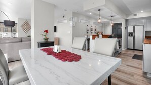 A modern kitchen with stainless steel appliances, a dining area with a marble table, and a living room with a gray sofa.