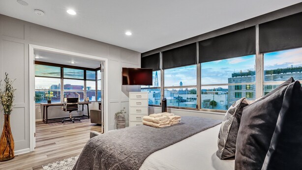A modern bedroom with a large bed, a desk, and a view of the cityscape through large windows.