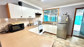 A kitchen with white cabinets, a black microwave, a stainless steel refrigerator, and a window with a view of a blue building. - Sunshine on Heath -Pet Friendly (Evans Head)