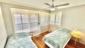 Una habitación con dos camas, un ventilador de techo, una ventana con persianas, una mesita con lámpara y una lámpara de pie.