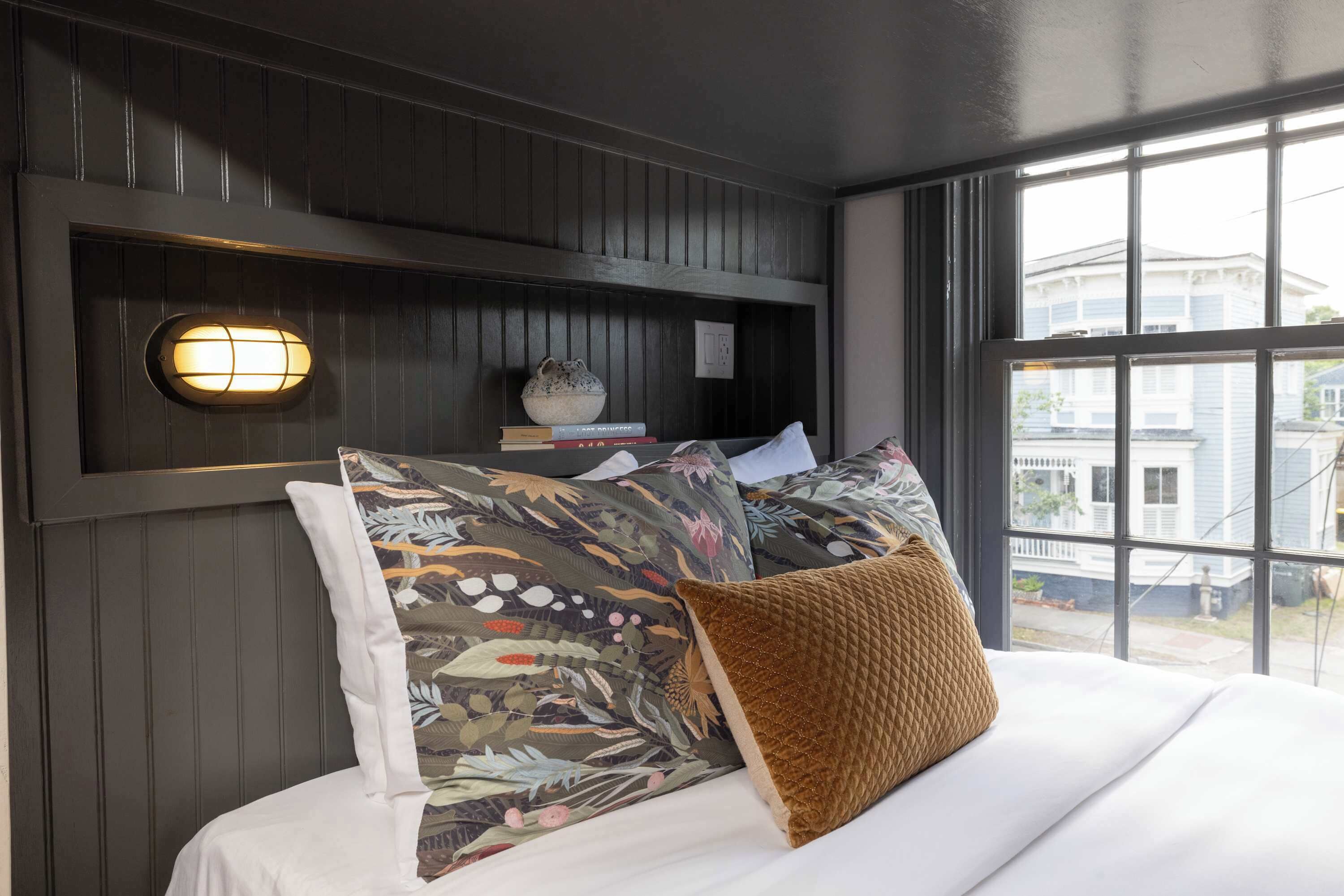 Ein Schlafzimmer mit einem Bett, einem Nachttisch, einer Wandlampe und Blick ins Freie durch ein Fenster.
