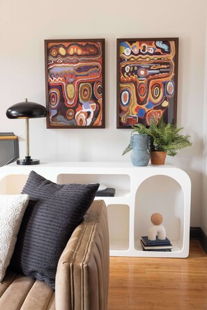 A modern living room with a sofa, a white console table, abstract paintings, a floor lamp, a potted plant, and a decorative item on the console table.