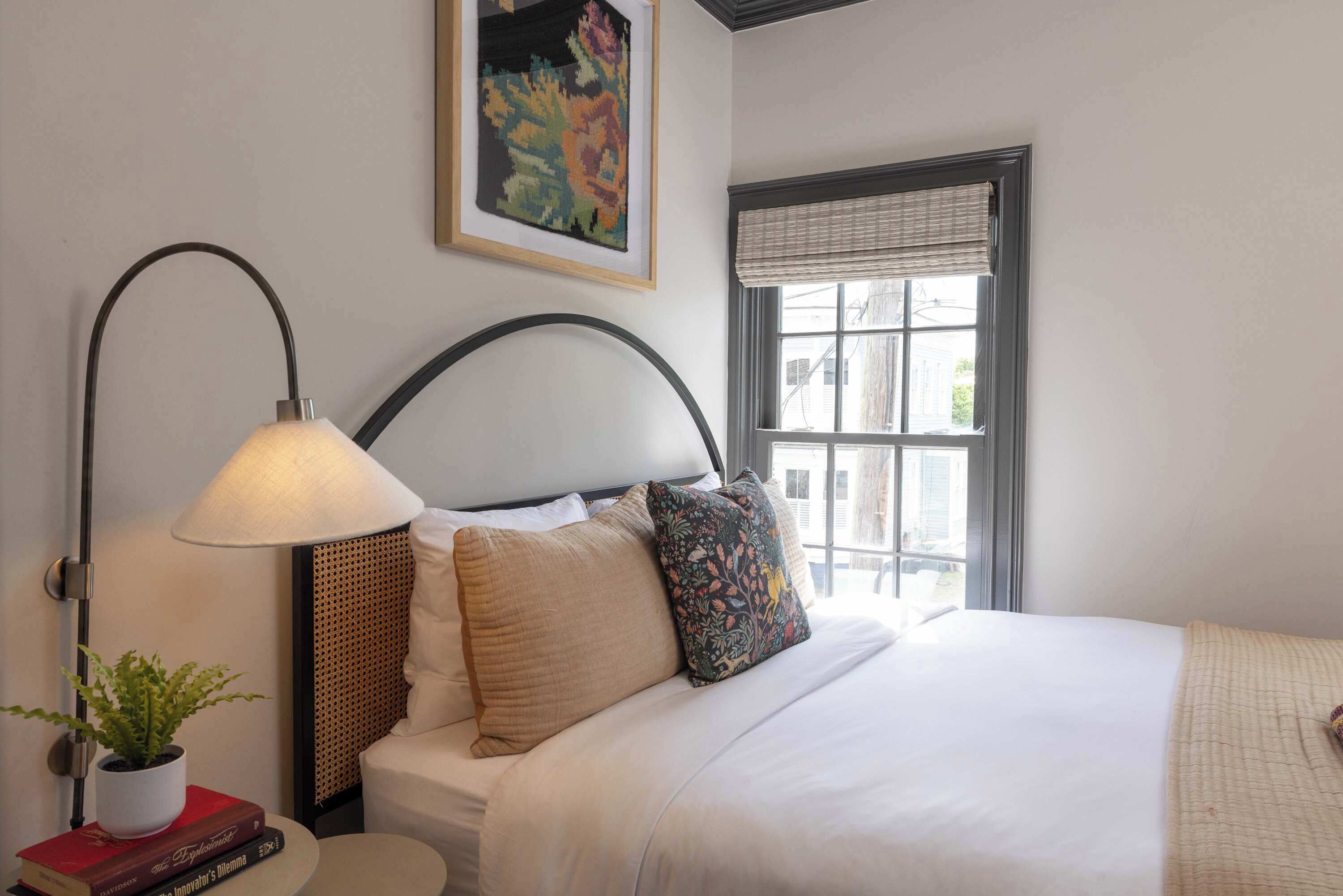 Un letto rifatto con un copriletto bianco e due cuscini, una lampada da comodino, una piccola pianta in vaso e un'opera d'arte incorniciata sopra il letto.