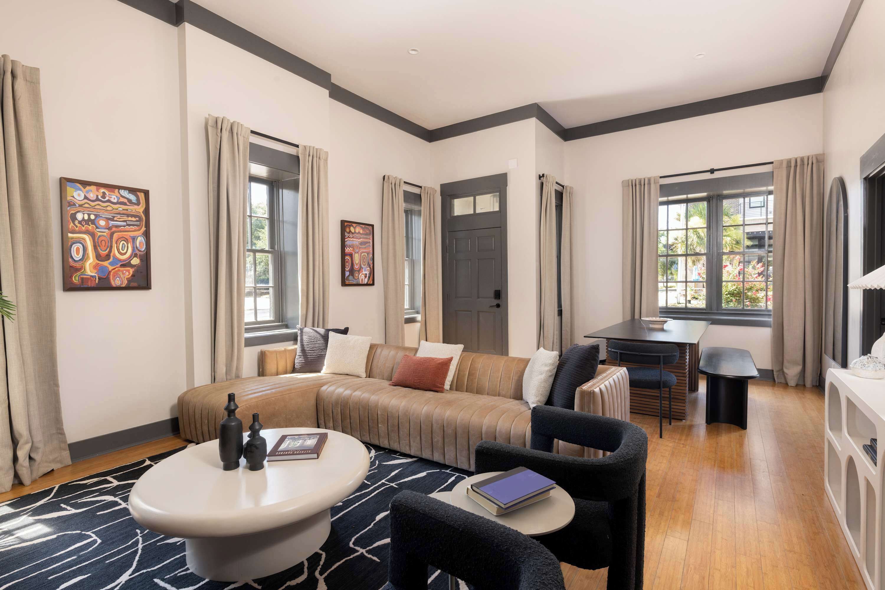 A modern living room with a sofa, coffee table, and chairs. There are windows with curtains, a door, and artwork on the walls.