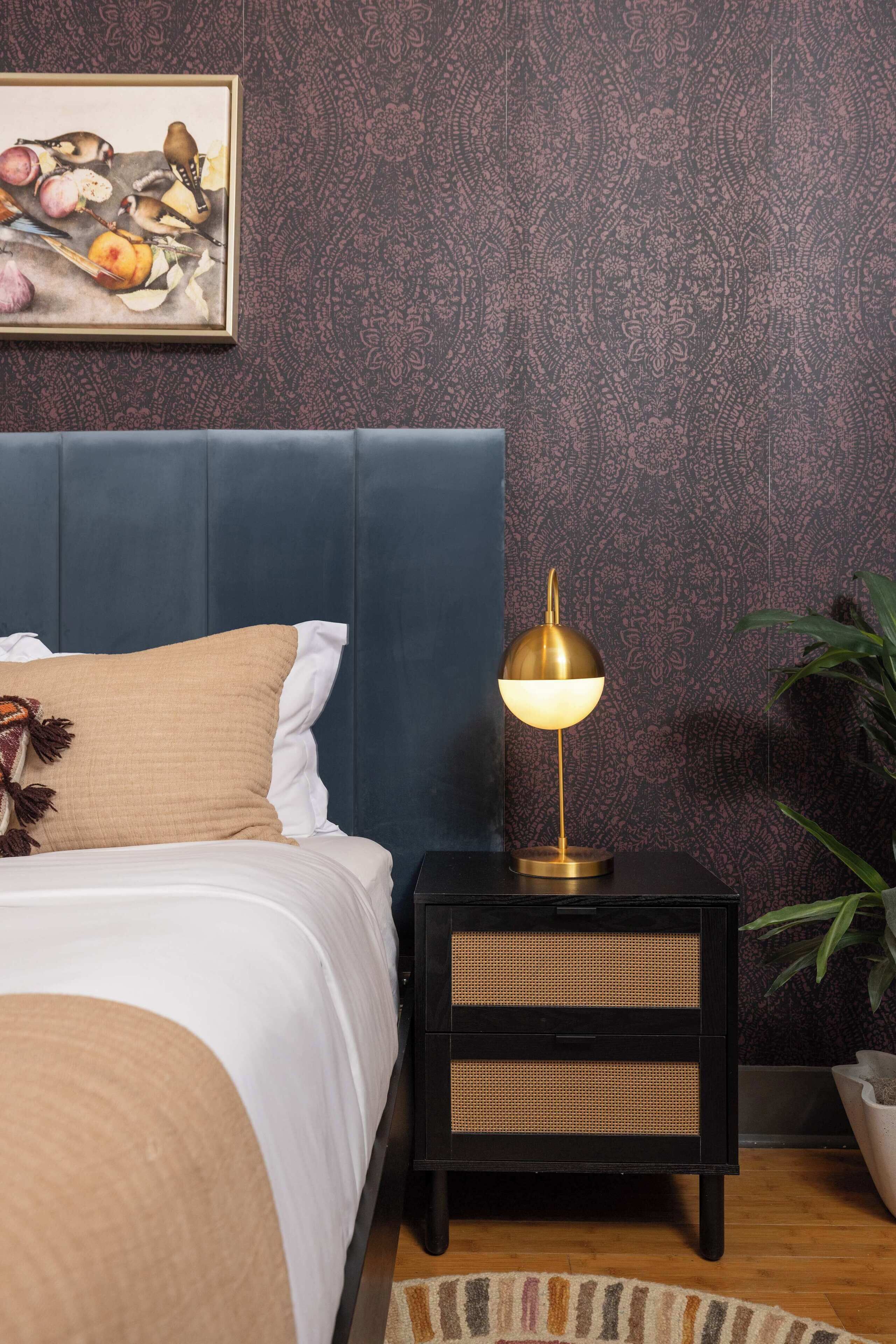 A bedroom with a blue headboard, a bedside table with a lamp, a rattan drawer, and a wall-mounted painting.