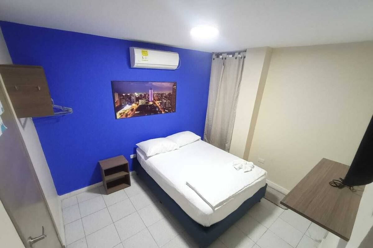 A hotel room with a bed, a small bedside table, a wall-mounted air conditioner, a picture on the wall, and a curtain.