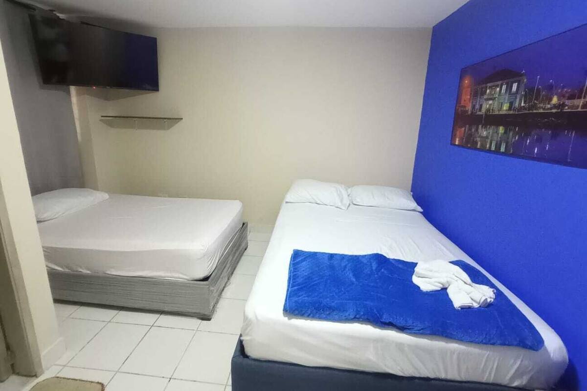 A small, compact room with two single beds, a flat-screen TV mounted on the wall, and a painting of a cityscape.