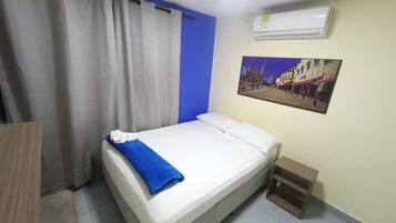 A hotel room with a bed, a small wooden nightstand, a wall-mounted air conditioner, and a painting of a building.