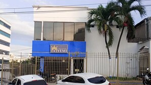 Hotel genaamd "Casa Hotel Yemi" met een blauw bord, twee witte auto's geparkeerd voor de deur, en een metalen poort.